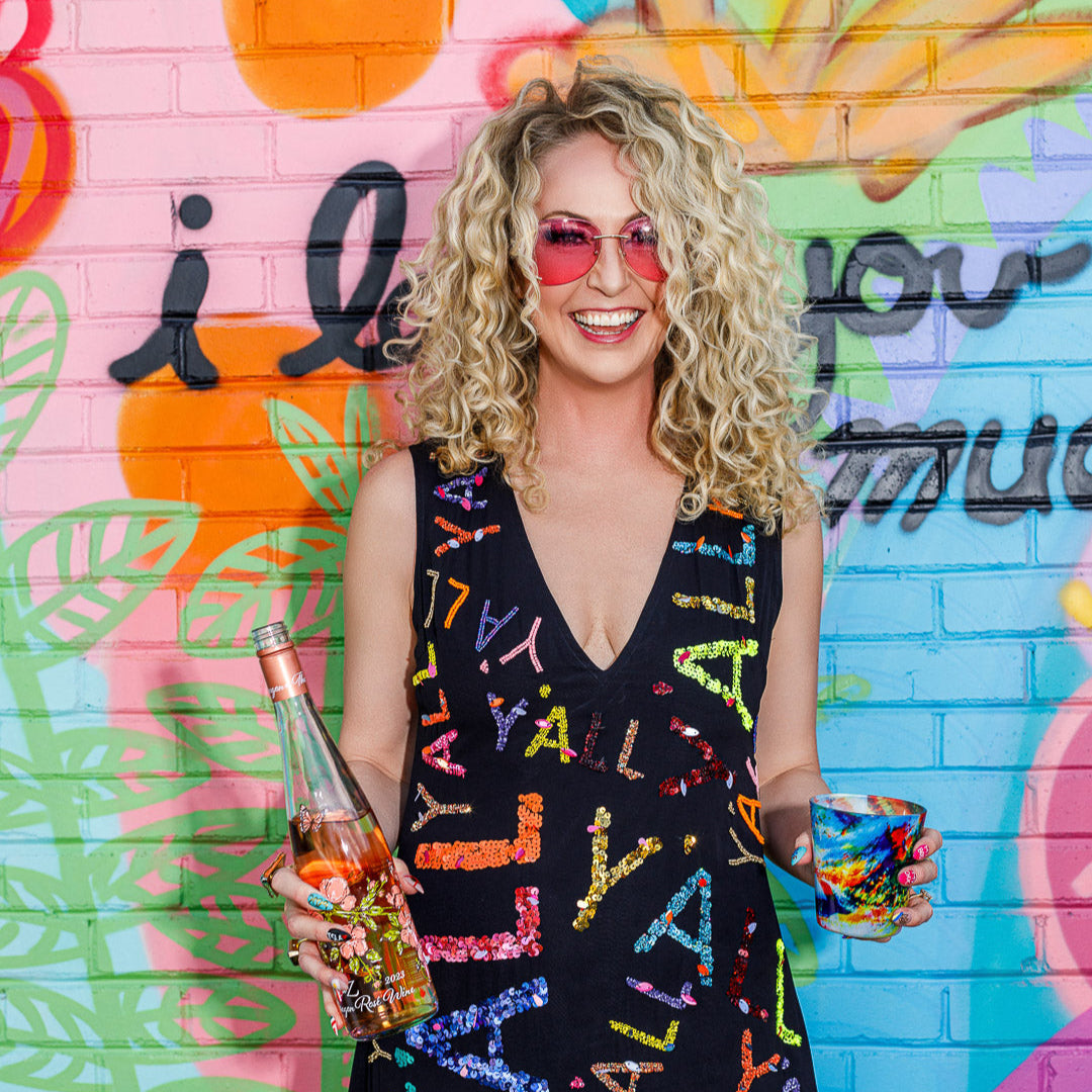 Woman in a colorful dress with 'Yall' text, holding drinks against a vibrant graffiti wall.