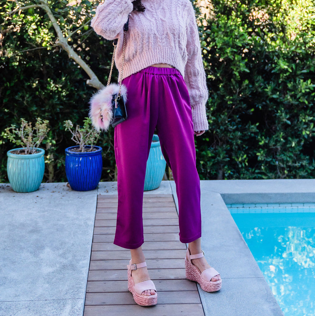 Woman in pink sweater and purple pants standing by a pool with greenery in the background