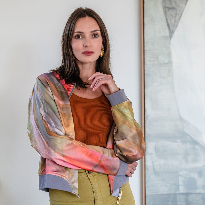 Woman wearing a colorful jacket over a brown top and green pants, standing in front of a painting.