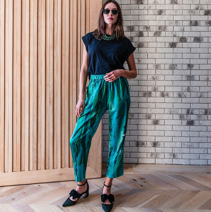 Woman in black top and green pants standing in a room with wooden paneling and brick wall.