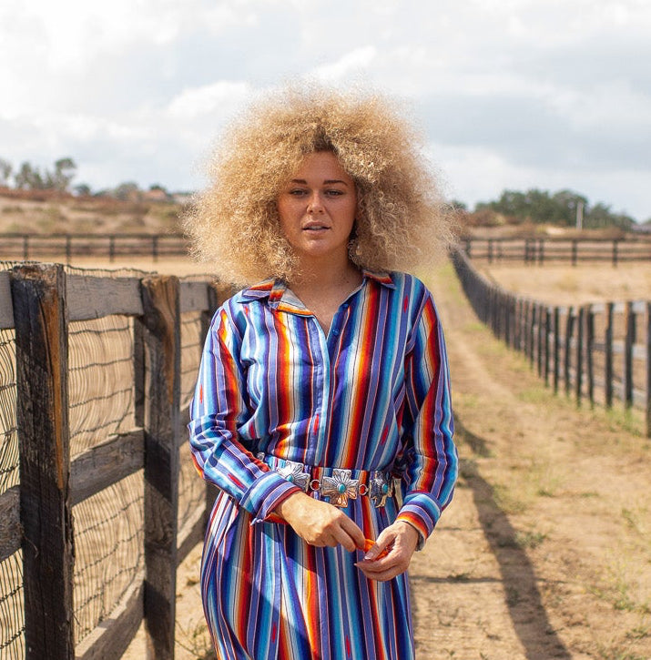 A woman with a large blonde wig stands in a dirt paddock next to a wooden fence, wearing a long-sleeve maxi dress with vertical red, blue, and white stripes. A horse is visible behind the fence on the left.