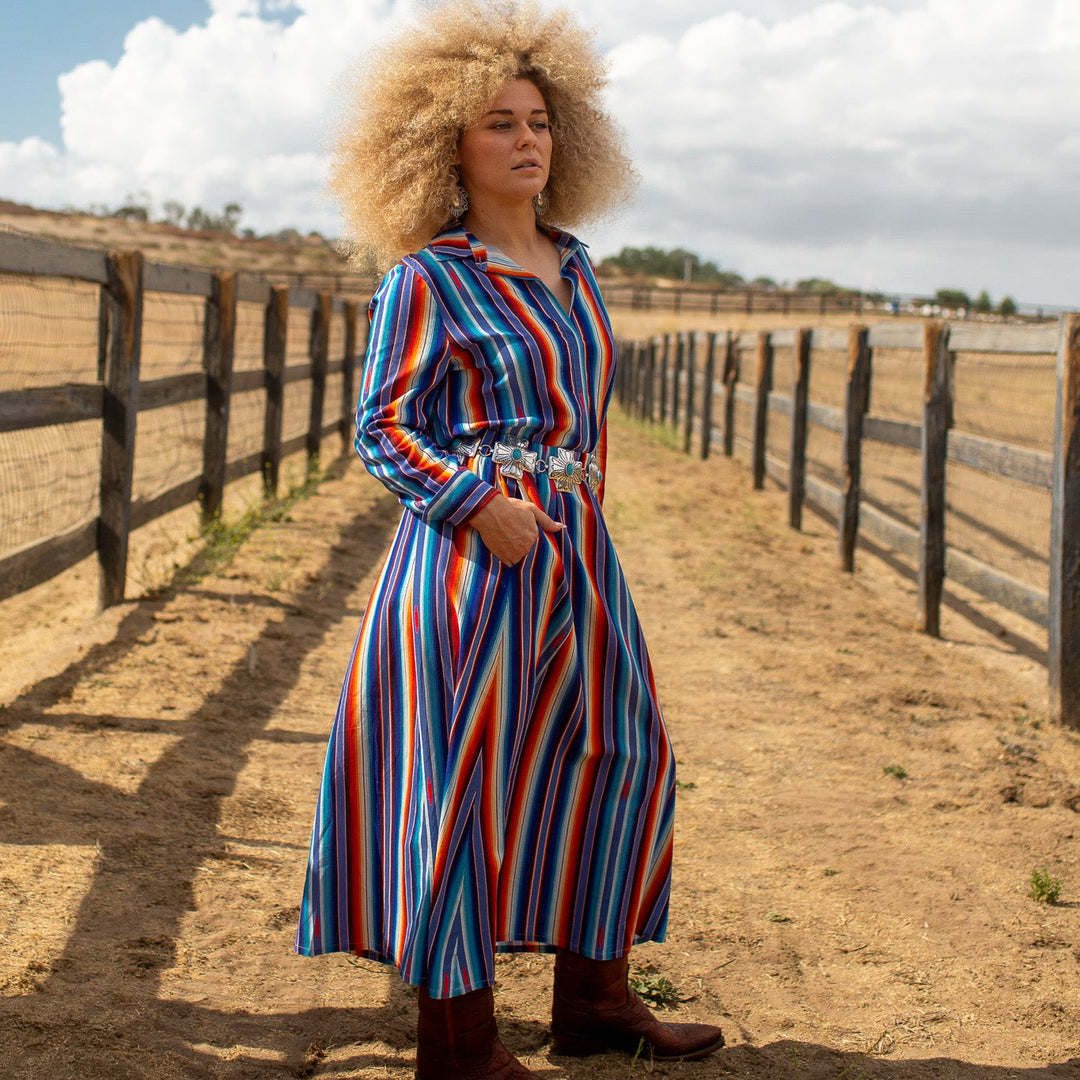 A woman with a large blonde afro stands outdoors on a dirt path between wooden fences under a blue sky, wearing a long-sleeve midi dress with vertical red, blue, and white stripes, a white belt, and brown boots.