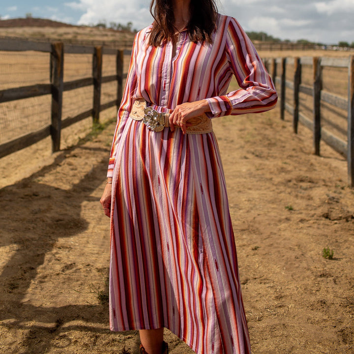 A woman wearing a long-sleeve red and white striped midi dress, a beige cowboy hat, sunglasses, and a decorative belt stands outdoors in a sunny, fenced field under a blue sky.