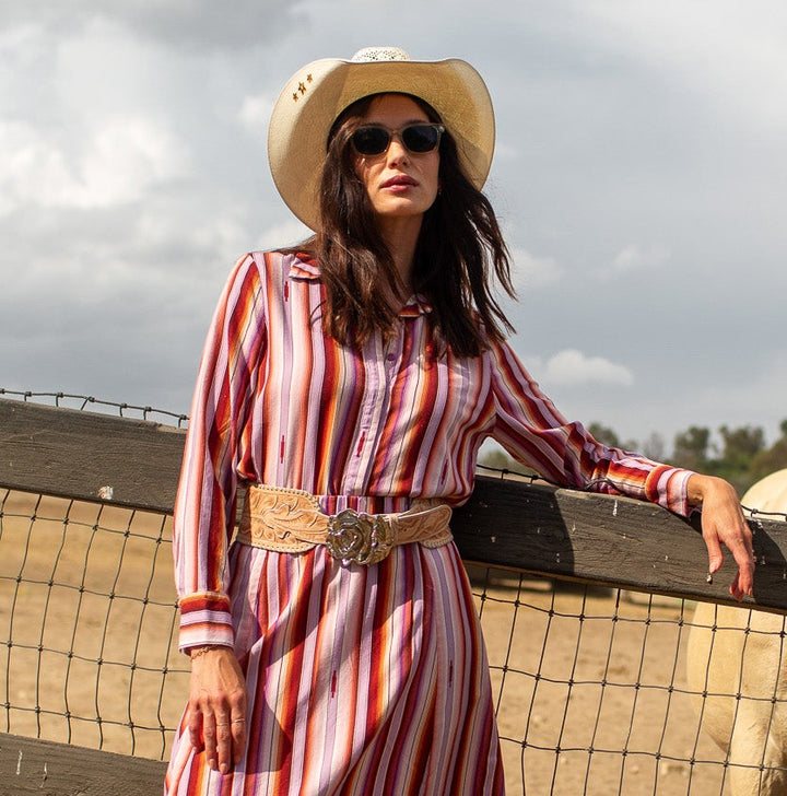 A woman wearing a red and white vertically striped long-sleeve midi dress with a matching belt and cowboy hat stands next to a wooden fence in an outdoor ranch setting, with a white horse behind the fence.