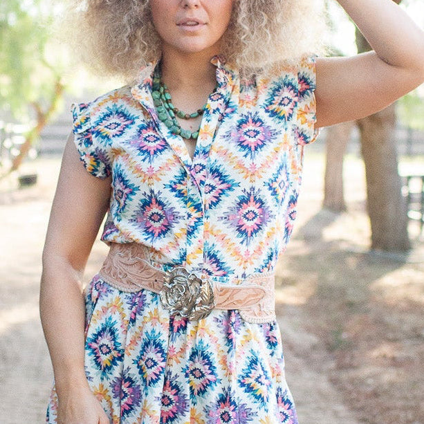 A woman stands on a dirt path outdoors wearing a sleeveless bohemian-style maxi dress with a colorful geometric diamond pattern, a matching belt, and metallic boots.