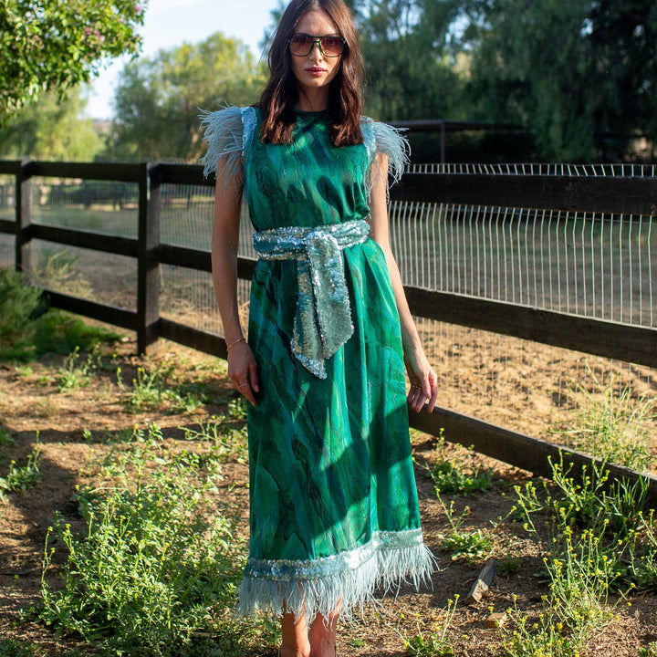 A woman wearing a green fringed midi dress with a matching belt and sunglasses, standing outdoors in a sunny field with a wooden fence and trees in the background.