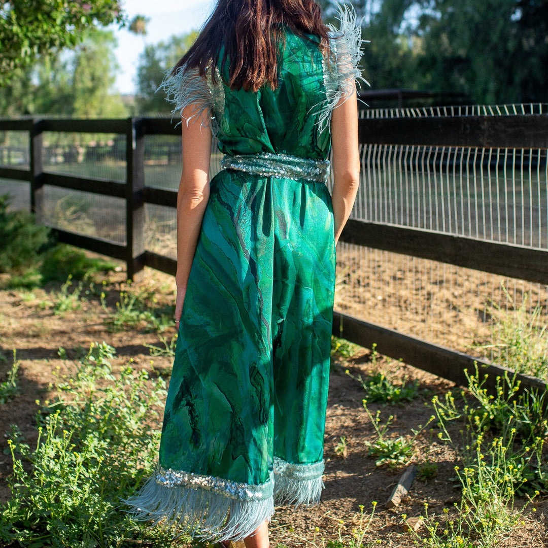 A woman viewed from behind wearing a green bohemian-style maxi dress with silver fringe and a silver waistband, standing outdoors in a garden next to a wooden fence.