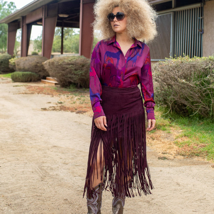 A woman stands outdoors on a dirt path wearing a long-sleeve collared shirt with a purple and blue paisley pattern, a dark burgundy fringe skirt, and brown cowboy boots. She has an oversized blonde wig and sunglasses.