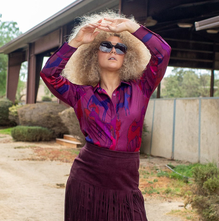 A woman posing outdoors in a vibrant purple and magenta patterned long-sleeve shirt, a dark purple fringe skirt, and brown cowboy boots, standing on a dirt path in front of a rustic barn.