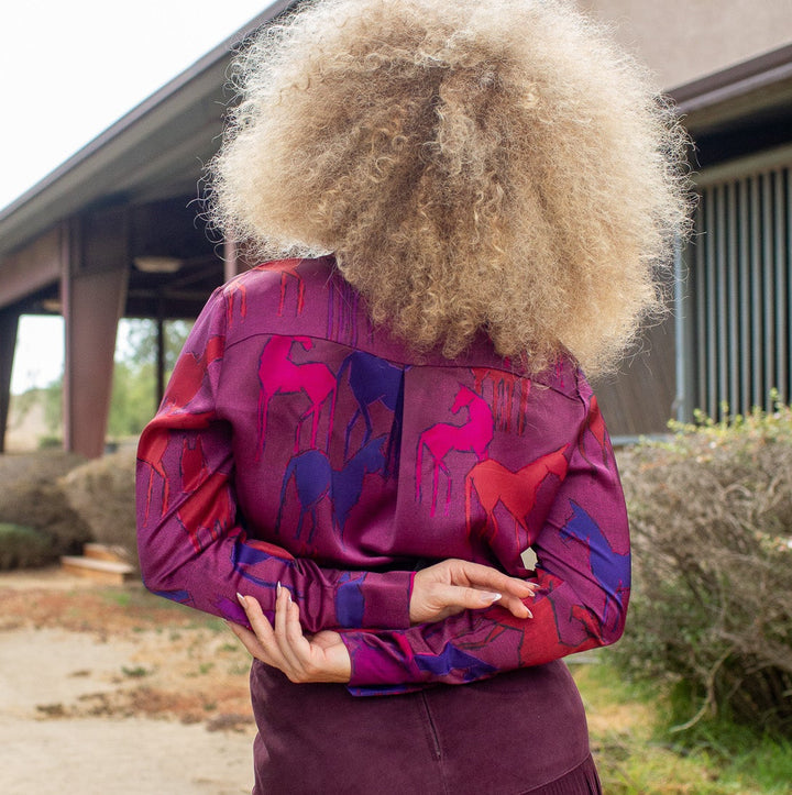 A person standing outdoors with their back to the camera, wearing a voluminous blonde afro, a long-sleeve patterned shirt in purple, pink, and blue, and a dark purple fringe skirt.