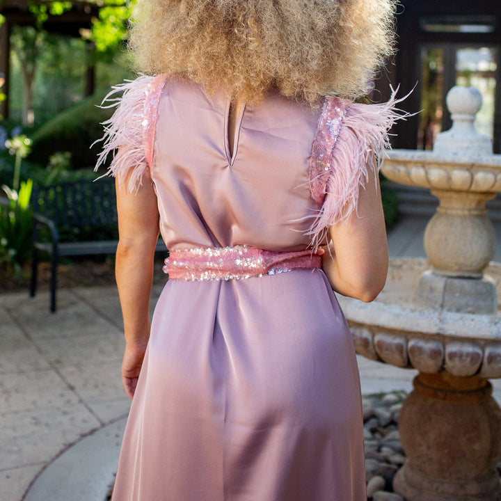 A woman viewed from behind wearing a lavender pink feathered gown in an outdoor garden setting with a stone fountain.