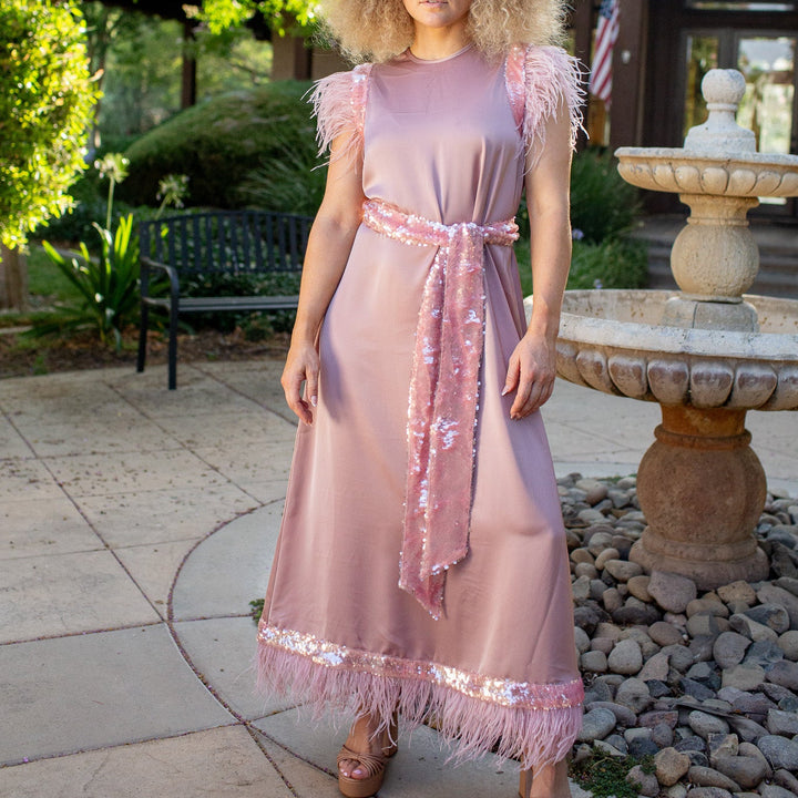 A woman stands outdoors in a garden courtyard wearing a dusty pink maxi dress with feathered cap sleeves, a matching sequined waist tie, and fringe trim at the hem. She is standing next to a stone fountain and trees.