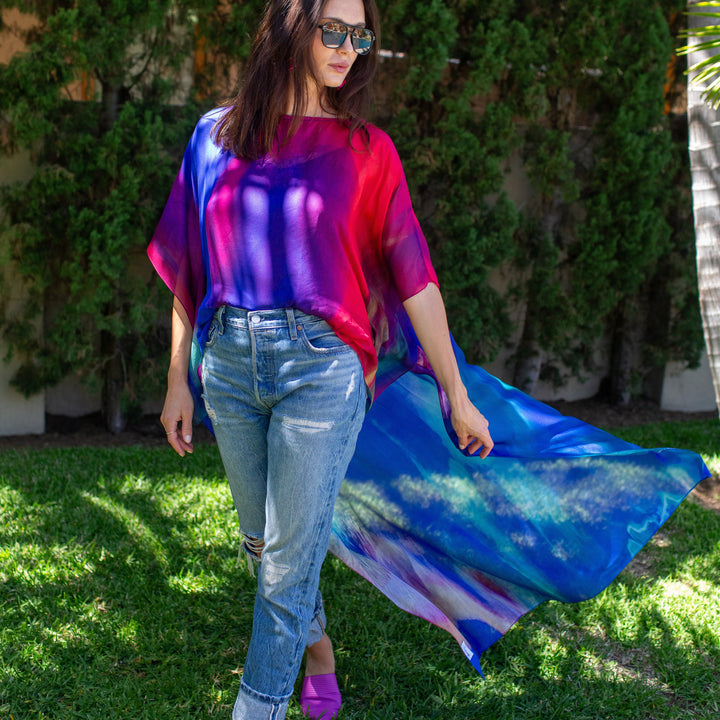 A woman wearing a vibrant tie-dye tunic top with blue jeans and purple pointed shoes, walking outdoors on a grassy lawn with a lush green hedge in the background.