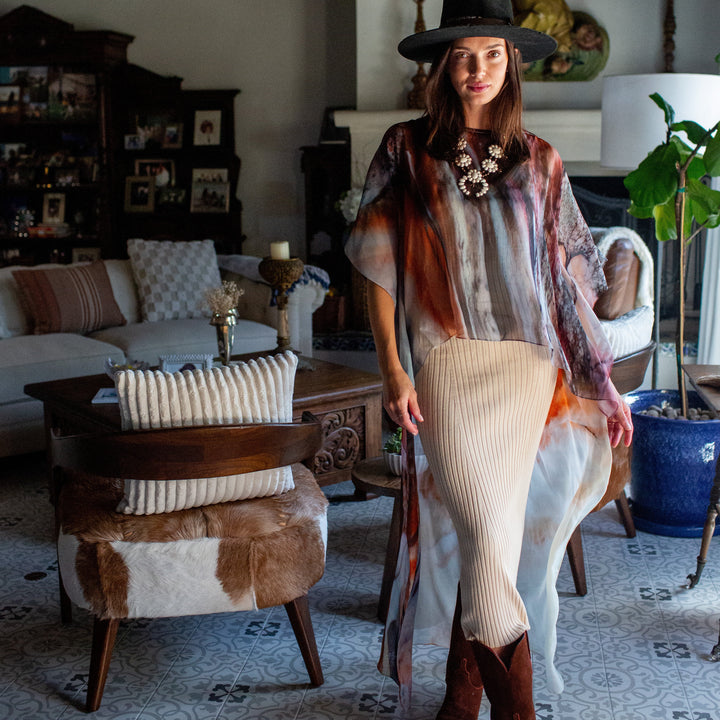 A woman wearing a bohemian-style outfit with a black wide-brim hat, a multicolor abstract print top, a cream ribbed skirt, and brown boots, standing in a decorated living room.