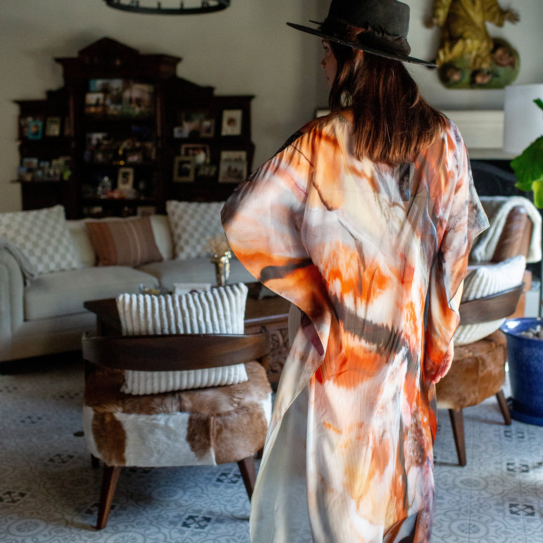 A woman wearing a wide-brim black hat and a flowing orange, white, and brown abstract-print kimono robe stands with her back to the camera in a rustic, bohemian-style living room.