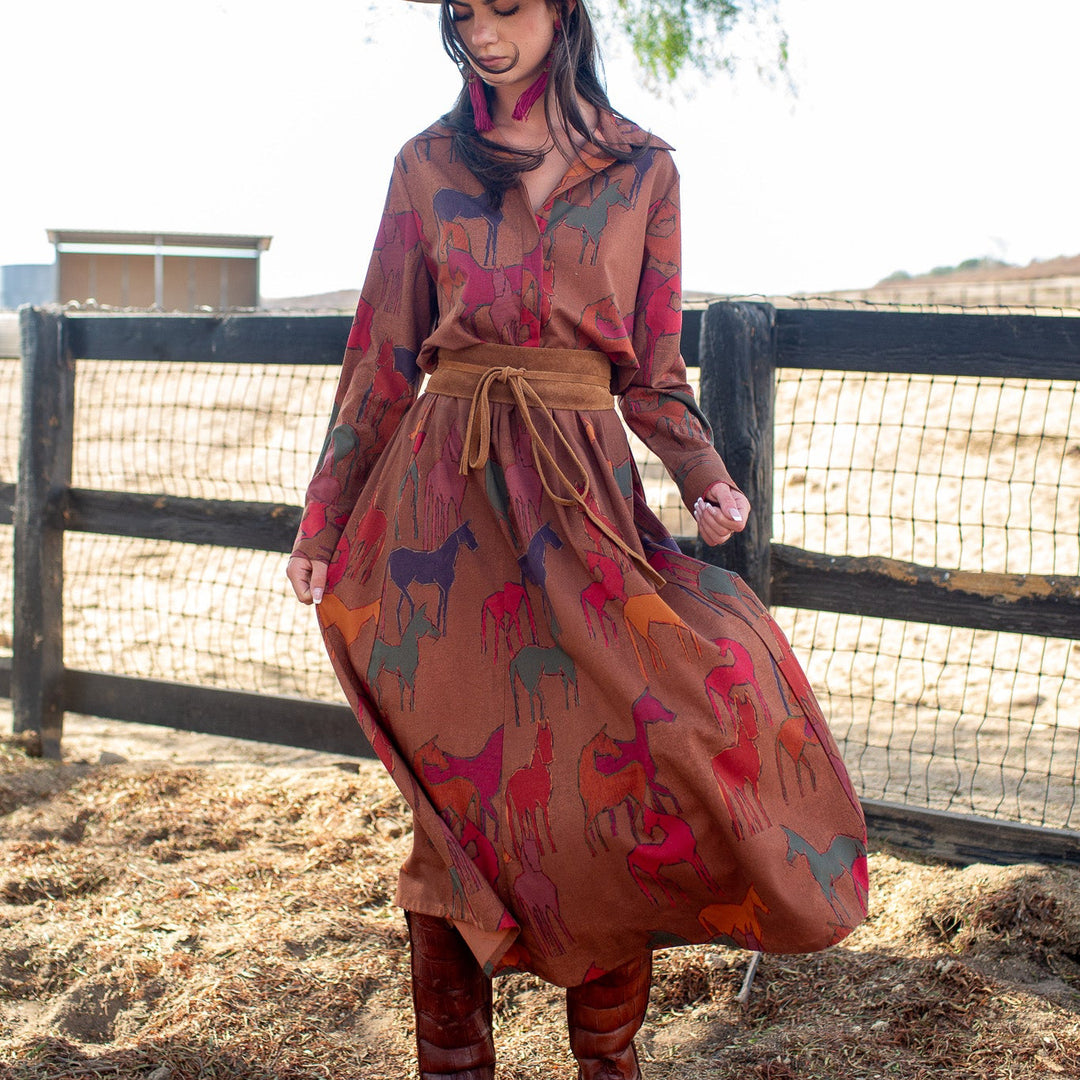 A woman wearing a long-sleeve rust-brown dress with a colorful horse print, a brown wide-brim hat, and brown boots, standing outdoors in front of a wooden fence.