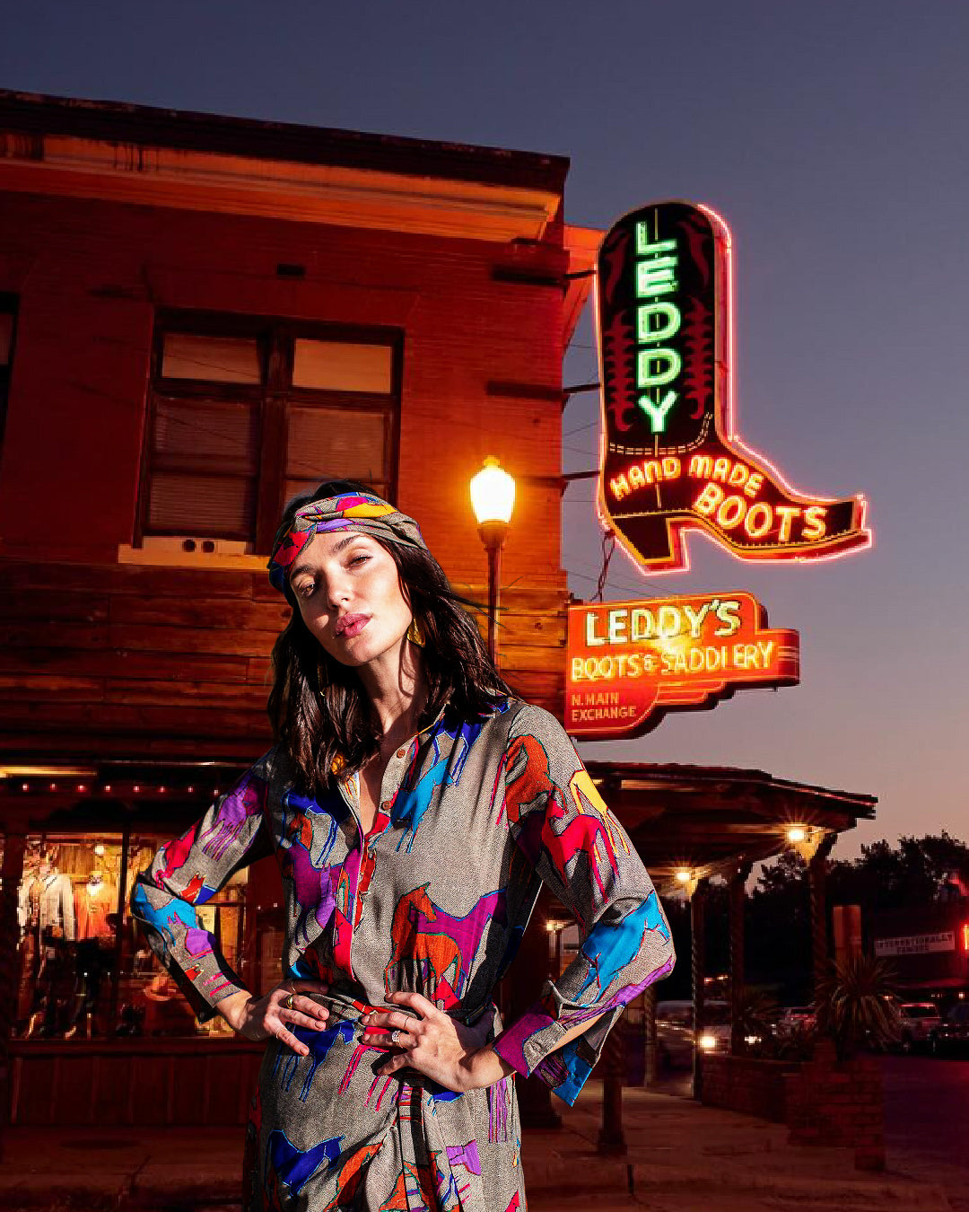 Woman in a colorful outfit standing in front of a neon sign for Leddy's Hand Made Boots.