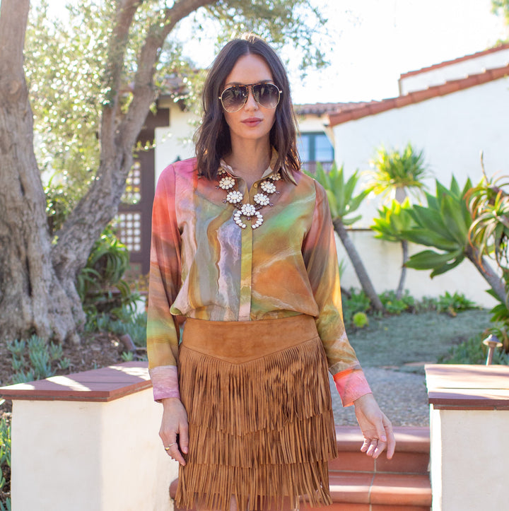 Woman in colorful top and fringed skirt standing outdoors with trees and a building in the background