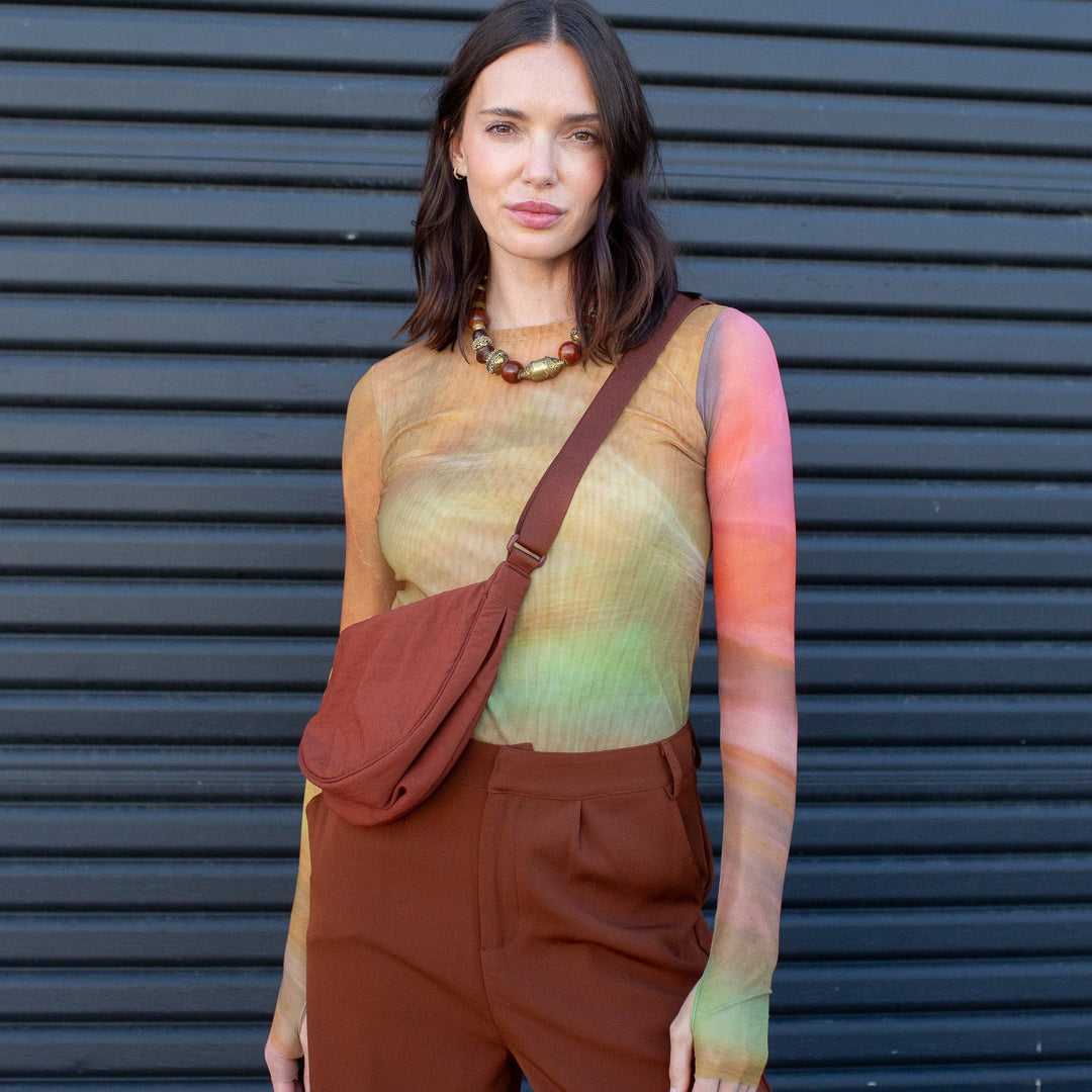 Woman wearing a colorful long sleeve top and brown pants with a red bag against a dark background